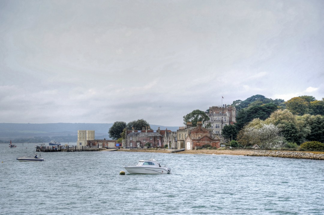 A view across the harbour towards Poole's iconic Brownsea Island, with boats dotted along the waterline. A prominent building composed of traditional red and grey brick sits in the background